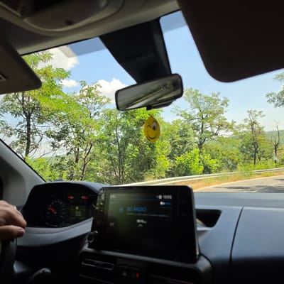 The image is taken from inside a car, looking out through the windshield. A hand with a ring is visible on the steering wheel. The dashboard displays a speedometer reading of 55 and a car radio screen showing "BG RADIO". A yellow smiley face air freshener hangs from the rearview mirror. The exterior view shows a road curving through a densely forested area with green trees and a blue sky with some clouds. The scene appears to be a road trip in the vicinity of Bratsigovo, Bulgaria, likely during the daytime with clear weather.