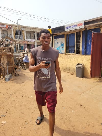 The image depicts an outdoor scene in what is identified as the city of Candelaria, Colombia, under clear daylight conditions. A young adult male, with dark skin and short, dark hair, is the primary subject, positioned slightly left of center and walking towards the viewer's right. He wears a dark brown sleeveless t-shirt featuring a light-colored graphic print that includes Chinese characters, and maroon cargo shorts with zippered thigh pockets. He is wearing grey slide sandals. His left hand holds a black smartphone, while his right hand is slightly open at his side. His gaze is directed forward and slightly downwards.

The ground is unpaved, consisting of light brown, sandy earth with scattered debris. In the background on the left, a stack of wooden structures or crates is visible, behind which a partially obscured individual in blue attire is seated. Further back, a two-story building with multiple windows and balconies is present. Overhead, utility lines traverse the sky, and a streetlamp is visible.

On the right side of the background, a single-story commercial building with light yellow walls features a prominent sign reading "HEALTHY CHOICE DOMI" with smaller text below it. The building has multiple windows with blue-tinted glass, and a white and blue poster is affixed to one window. A grey outdoor waste bin is positioned to the right of the windows, adjacent to a red metallic panel or door. The sky is bright and appears mostly clear.