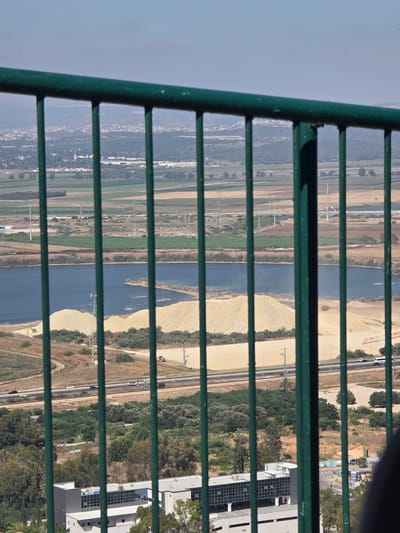 The image is a tilted, vertical shot looking down through a green metal railing. The scene is a wide landscape of a river, agricultural fields, a highway, and some buildings, seen from an elevated perspective. The river is a prominent blue feature, with sandy banks and vegetation on either side. Beyond the river, there are fields with varying shades of green and brown, suggesting different crops or stages of growth. A highway with cars is visible, running parallel to the river. In the background, there are some modern buildings with glass facades, one of which appears to be an industrial or commercial complex. There are also trees and some power lines or towers in the landscape. The time of day appears to be daytime, with clear skies, indicated by the bright sunlight and shadows cast by the railing. The overall impression is of an arid or semi-arid region with evidence of human activity and infrastructure interspersed with natural and agricultural elements. The location is identified as Nesher, Israel.