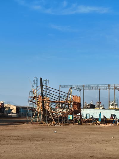 This media file depicts a ship construction site in Chimbote, Peru. The scene is dominated by the skeletal structure of a ship in progress, supported by a complex network of scaffolding. Several individuals, likely workers, are visible at various points around the construction, engaged in what appears to be ongoing work.
The setting is an open, dusty area, typical of a shipyard. In the background, other structures and possibly other vessels under construction or repair can be seen, along with a long, light blue building. The sky is a clear, vibrant blue with a few wispy clouds, suggesting fair weather. The lighting indicates it is daytime, likely morning or afternoon, given the bright sunlight casting shadows.
Notable details include the intricate scaffolding made of metal poles and wooden planks, forming the framework for the ship's hull. There's a yellow caution sign with a symbol of a person falling, indicating safety awareness on site. The ground is rough and unpaved, with tire tracks visible in places. The presence of boats in the background, combined with the ship construction, strongly suggests a maritime or shipbuilding industry setting. The overall atmosphere appears to be one of active work and industrial activity.