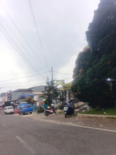 The image depicts an overcast street scene, likely in a town or city. Visible text elements within the image, such as "Kec. Pagerageung" and "Griya Prama," are Indonesian in origin, indicating the depicted location is in Indonesia and not Morrumbala, Mozambique, as specified.

The sky is uniformly overcast and light grey, occupying the upper half of the frame. Multiple overhead power lines extend across the upper left and central portions, supported by at least one visible utility pole.

An asphalt road runs horizontally through the lower half of the image. On the left side of the road, a small white van and a blue minivan/truck are parked on the shoulder, facing away from the viewer. Behind these vehicles, a row of one-to-two-story buildings, appearing to be shops or residences, lines the street. Signage, including a blue banner displaying "Kec. Pagerageung," is visible on these structures.

On the right side of the road, a dirt and gravel shoulder transitions to a slightly raised unpaved area. Two dark-colored scooters are parked here. The first scooter, positioned slightly forward, faces left and has a yellow object on its seat. The second scooter, further back and closer to a dark metal gate, has one person seated upon it, wearing a dark top and a helmet. This individual appears stationary. Above the gate, a green sign, partially obscured, reads "Griya Prama." A large, dense tree with dark green foliage and scattered reddish-orange elements dominates the right side of the image, extending upward from mid-frame. Smaller trees and vegetation are also present along the roadside.
