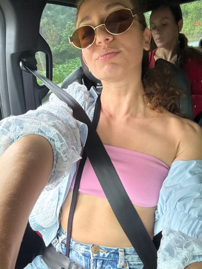The image captures two women inside a vehicle, located in Dế Xu Phình, Vietnam. The woman in the foreground is taking a selfie, her head tilted slightly downward. She wears gold-framed, brown-lensed sunglasses, a pink ribbed strapless tube top, and light blue acid-wash denim jeans. A light blue garment with white lace-trimmed sleeves is worn over her top, appearing open or draped. A black seatbelt is fastened across her chest and lap. Her right arm, covered by the light blue sleeve, is visible on the left side of the frame. Her brown, curly hair is visible over her right shoulder. In the background, a second woman is partially visible behind the foreground subject. She has dark hair pulled back and is wearing a red garment. The vehicle interior features black components, including the B-pillar and seatbelt hardware. Through the windows, a lush green landscape of foliage and what appears to be vegetation is visible. A yellow triangular road sign with an indistinguishable black symbol is faintly discernible in the far background.