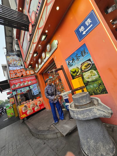 The image shows a street scene in Gongshu District, China, featuring a brightly colored food establishment with an orange facade. A woman in a blue patterned jacket and blue pants stands near the entrance, holding a rope attached to what appears to be a traditional grinding stone. In front of the establishment, several food stalls display menus with images of various dishes, including dumplings and soups. A blue sign with white lettering points to the "Subway" (地铁). The street is paved with gray tiles, and a stone curb separates the sidewalk from the road. The overall impression is a lively urban setting with a focus on food vendors.