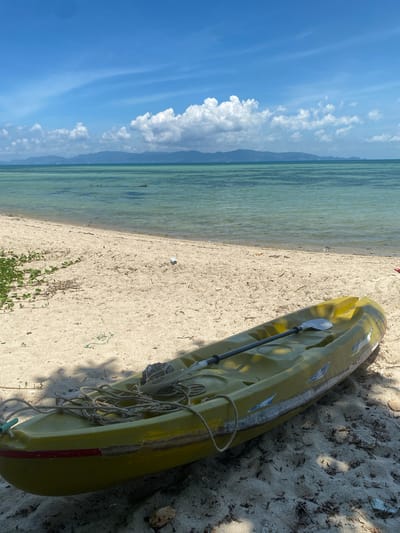 This image depicts a serene tropical beach scene in Moo 1, Thailand, under a clear, bright blue sky dotted with fluffy white clouds. The photograph is oriented vertically, showcasing a sandy shore, the clear turquoise sea, and distant land.

In the foreground, a lime green and yellow kayak rests on the light-colored sand, angled towards the water. A paddle is laid across the top of the kayak, and several coils of rope are gathered near its stern. The kayak shows signs of wear, with visible scratches and patches, and a red stripe near its tail. Shadows cast by the kayak and unseen foliage suggest bright sunshine, likely late morning or early afternoon. Small green plants and some scattered debris are visible on the sand near the water's edge and around the kayak.

The calm, shallow sea stretches across the midground, exhibiting clear, translucent waters with shades of green and blue. In the background, a low-lying range of mountains or distant land mass is visible across the horizon, partly obscured by the cloud formation. No people are present, contributing to the tranquil and undisturbed atmosphere of the beach. There is no visible text in the image.