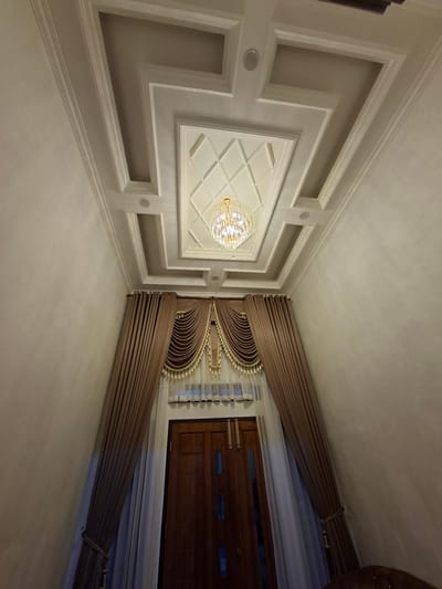 The image displays an interior view of a room, likely a living room or entryway, in Uleemeuria, Indonesia. The focal point is the ornate ceiling, which features a multi-layered, coffered design with recessed lighting and a crystal chandelier hanging from the center. The ceiling is painted in shades of white and grey, with intricate molding details.
Dominating the lower half of the image are heavy, brown velvet curtains that frame a wooden door and a sheer white curtain. The door appears to be a double door, made of dark wood, and has visible rectangular glass panels. The curtains are draped and adorned with gold tassels and a decorative pelmet. The walls are painted a neutral, textured beige. The overall impression is one of a luxurious and well-decorated interior. There are no people visible in the image.