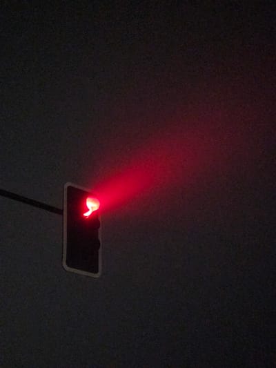In San Fernando, Chile, a traffic light shines a bright red, casting an illuminated beam into the dark, overcast sky. The traffic light is black and rectangular, with a single red light illuminated, emitting a strong glow that stretches towards the right of the frame. A thin, dark horizontal line, likely a support cable, is visible on the left side of the image, intersecting the frame. The background is a uniformly dark and slightly textured sky.