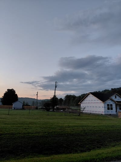 The image depicts a partially constructed house, wrapped in white protective sheeting with "Builders" branding, situated in a grassy field. The house has a dark roof and white-framed windows. In the background, a line of trees forms a backdrop against a sky with scattered clouds, transitioning from a pale blue to a softer hue near the horizon. A small, light-colored shed with a dark roof is visible to the left, behind a wooden fence. Utility poles with wires are also present in the scene. The foreground is dominated by a well-maintained green lawn, with visible rows suggesting recent mowing. The overall impression is of a rural or semi-rural setting, possibly a suburban or exurban area, at dusk or dawn. The exact location in Jeffersonville, United States of America, cannot be determined from the image alone.