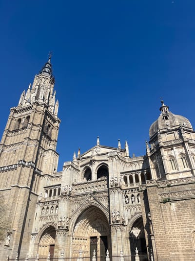 An upward-angled, daylight photograph captures the Primate Cathedral of Saint Mary of Toledo, commonly known as Toledo Cathedral, located in Toledo, Spain. The monumental Gothic structure dominates the lower and central portions of the frame against a clear, deep blue sky, which occupies the upper half.
On the left, the cathedral's tall, intricately detailed North Tower rises prominently. It transitions from a robust square base to an octagonal spire adorned with multiple pinnacles, lancet windows, and decorative elements, culminating in a pointed finial topped with a cross and a weather vane. The tower exhibits light-colored, weathered stone.
The central facade features multiple arched portals, deep recesses, and numerous niches containing carved statues. Intricate Gothic tracery, balustrades, and arcaded galleries are visible across its upper sections, primarily constructed from a lighter, more ornate stone. Shadows from direct sunlight delineate architectural features, particularly on the lower and central portions of the facade. A dark metal fence with decorative finials runs across the bottom foreground, partially obscured by minimal foliage from a bare tree branch on the far left.
To the right of the central facade, a prominent, grey stone dome (cupola) rises, featuring arched windows and ornate detailing, capped with a finial and cross. This structure is integrated into the broader cathedral complex.
The sky in the upper right quadrant reveals a faint, illuminated crescent moon. No individuals are visible within the frame. The scene is static, devoid of discernible actions or interactions.