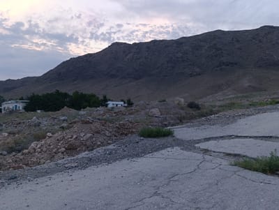 The image shows a rugged, mountainous landscape with a cracked asphalt road in the foreground. The sky is overcast with scattered clouds. In the middle ground, there are some buildings and trees, suggesting a settlement. The overall impression is one of a remote and somewhat desolate location. The presence of cracked roads and the natural terrain suggest a location in a rural or mountainous area. Given that Zarafshan City is known for its mining operations and is located in a desert region with surrounding mountains, this image could depict a road leading to or from such an area. The environment appears arid with sparse vegetation. There are no people visible in the image.