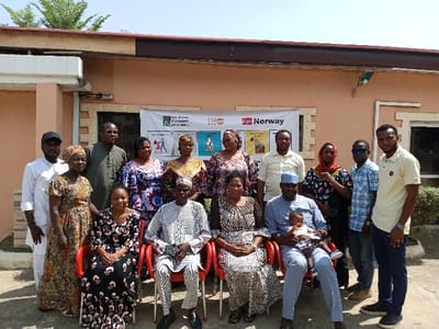 A group of 13 people stand and sit in front of a building in Yola, Nigeria, at the coordinates 9.259 latitude and 12.466 longitude. They are positioned in front of a large sign with the word "Norway" and the Norwegian flag. The sign is on the side of a peach-colored building with a concrete walkway in front of it. The people are all dressed in various styles of clothing. Some of them are smiling and others have serious expressions. Four people are seated in red chairs, and one person holds a small baby. The people are diverse, with varying skin tones and hair colors. There is a small amount of vegetation in front of the building.