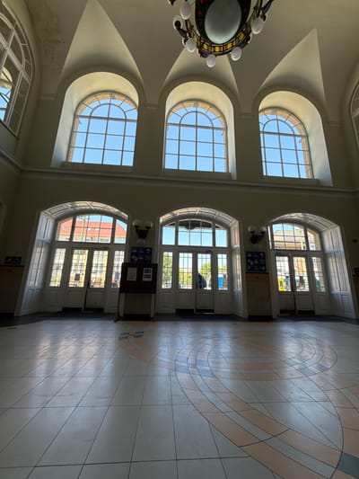 The image captures the interior of a large, grand public hall or station, located in Szeged, Hungary. The scene is dominated by classical architectural features, including high ceilings with coved arches, large arched windows, and equally grand arched doorways.
On the left side of the frame, a series of three tall, arched windows allow abundant natural light to flood the space. Through these windows, a clear blue sky is visible, along with glimpses of a brick building exterior. From the ceiling on the far left, a decorative chandelier with multiple bulbs and a central globe hangs.
On the right side, two large arched openings serve as main entrances or exits. These feature paneled white doors with glass sections, revealing views of distant buildings and trees outside. Between these two lower arched doorways, a dark brown information stand is positioned, featuring several blue directional signs with white pictograms and arrows. These signs clearly indicate directions for "train" (left arrow), "bus" (right arrow), "parking" (down arrow), and an icon for "lodging/hotel" (left arrow). A smaller blue sign with a right-pointing arrow and a stylized human figure is also visible near the floor on the lower left.
The floor is covered with light-colored tiles, featuring a prominent, large circular geometric pattern in shades of cream, beige, and light blue/grey. The bright natural light streaming in suggests it is daytime, likely morning or early afternoon, creating distinct areas of sunlight and shadow across the floor. No people are visible, lending a quiet and spacious feel to the scene. The overall impression is one of historical elegance combined with functional public utility.