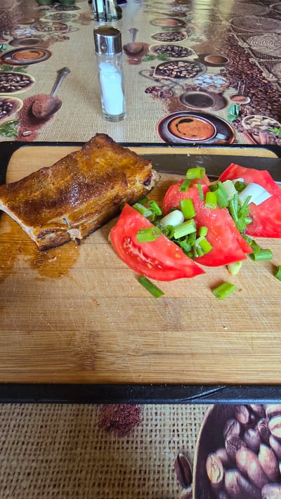 This image captures a meal served on a wooden cutting board, situated on a patterned tablecloth, within a domestic setting in Gorni Bogrov, Bulgaria. The primary subject is a substantial piece of cooked meat, likely a cut of ribs or belly, positioned on the left side of the cutting board. It exhibits a crispy, golden-brown exterior with visible layers of fat and is lightly seasoned with coarse white salt crystals. Adjacent to the meat, on the right side of the board, are multiple thick slices of red tomato, approximately three to four, topped with chopped green scallions and small white fragments, possibly white onion or garlic. Fine salt crystals are also visible on the vegetables.
The cutting board itself is light brown, rectangular, and shows significant signs of use, including numerous parallel and intersecting knife marks and scratches across its surface. In the upper-middle background, a clear glass salt shaker with a metallic silver-colored top stands upright, containing white granular salt. The cutting board rests upon a textured tablecloth patterned with various coffee-related motifs, including coffee beans, ground coffee, cups of brewed coffee, and serving spoons. The tablecloth's base color is a light beige or natural fabric tone, suggesting a woven material like burlap, with the coffee imagery rendered in shades of brown and dark earth tones. The scene is illuminated by an overhead or ambient light source, casting soft shadows.