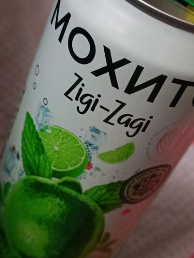 The image shows a close-up of a white can with black text and graphics. The text on the can reads "МОХИТО" and "Zigi-Zagi". There are graphics of lime slices, mint leaves, ice cubes, and red berries on the can. The can appears to be a beverage can, possibly for a mojito-flavored drink. The background is out of focus and appears to be a textured surface, possibly fabric. There is no indication of the location in the city of Гвардейский, Kazakhstan. The image is a still photograph and there are no people or actions depicted.