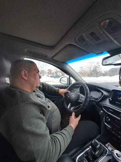 The image depicts the interior of a vehicle, identified as being in Новые Выселки, Russia, from a passenger-side perspective looking towards the driver. An adult male, with a closely shaved head and wearing a dark olive-green long-sleeved sweatshirt and black trousers, occupies the driver's seat. He is secured by a black seatbelt across his chest and lap. Both hands are placed on the steering wheel, which is fitted with a black cover, indicating an active state of driving or readiness to drive. The vehicle's interior features a grey headliner with an overhead console and a visible sunroof. The dashboard contains a digital instrument cluster and a central infotainment screen. A rearview mirror with a beaded ornament hangs from the windshield. Through the windshield and driver's side window, the exterior environment appears to be a winter or late autumn scene, characterized by snow-covered ground, leafless trees, and distant residential structures under an overcast sky.