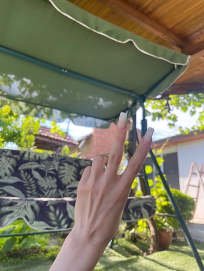 A first-person perspective shot captures a hand making a peace sign in front of a blurry outdoor scene. The hand, with long, square-shaped light-colored nails, is positioned centrally in the frame. Behind the hand, a green-canopied swing set is visible, with a wooden roof overhead. The background reveals a residential area with a tiled roof, trees, and a ladder leaning against a white structure. The setting appears to be a backyard or garden area in Totleben, Bulgaria, characterized by lush greenery and dappled sunlight.