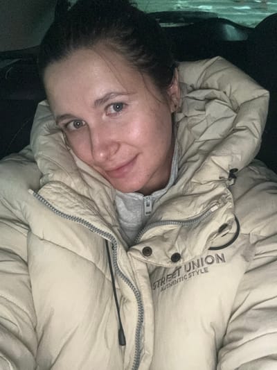 The image displays a close-up portrait of an adult female located in the city of Vorsha, Russia. She is situated within a dark, indistinct interior environment, consistent with a vehicle cabin, with blurred ambient light sources visible in the background. The female has dark hair, which is pulled back, light-colored eyes, and fair skin, exhibiting a slight smile while looking directly at the camera. She is wearing a light cream or beige puffer jacket. The jacket features a visible zipper, snap buttons, and a drawstring with a toggle. The text "STREET UNION AUTHENTIC STYLE" is printed on the wearer's right side of the jacket, accompanied by a partial circular logo. A grey zippered hoodie is visible underneath the puffer jacket.