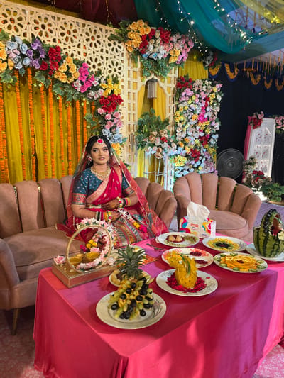A single adult female is seated on a light brown, plush upholstered sofa within a decorated indoor event space in Savar, Bangladesh. She is positioned towards the center-left of the frame, looking slightly to her right. The woman is adorned in traditional attire, consisting of a dark teal embroidered blouse, a fuchsia-red sari with gold trim, and a matching sheer veil over her head. She wears extensive gold-toned jewelry, including a large necklace, earrings, bangles on both wrists, and a maang tikka on her forehead. Henna patterns are visible on her hands.
In front of the woman, a rectangular table draped with a fuchsia pink tablecloth holds several items. These include a gold-colored tray with a circular mirror frame, containing two small yellow bowls. Multiple white plates are arranged on the table, displaying elaborately carved and arranged fresh fruits, such as sliced oranges, kiwis, grapes, pomegranate seeds, and pineapples (one carved into a decorative form). A large watermelon, carved into a basket shape and filled with various fruits, is positioned on the right side of the table. A white box of "PARTE X" brand facial tissues is also present.
The background features a decorative stage setup with vertical yellow fabric drapes and gold-colored lattice panels adorned with artificial floral arrangements in red, pink, yellow, blue, and white. An armchair matching the sofa is partially visible to the right. Overhead, green and yellow sheer fabric drapes are strung with fairy lights. A black stand fan is visible in the upper right background. The floor is covered with a light-colored, patterned carpet.