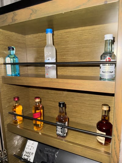 A liquor cabinet displays mini bottles. On the left, from top to bottom, are Bacardi rum, Grey Goose vodka, and an unlabeled blue bottle. On the right, a bottle of Hennessy rests on top, followed by Jack Daniels, a small bottle with a red stripe label, and an amber bottle. A black metal rod secures the bottles in each shelf. A white bag with “Hotels Worldwide, Take it away OMR 3” is visible in the background.