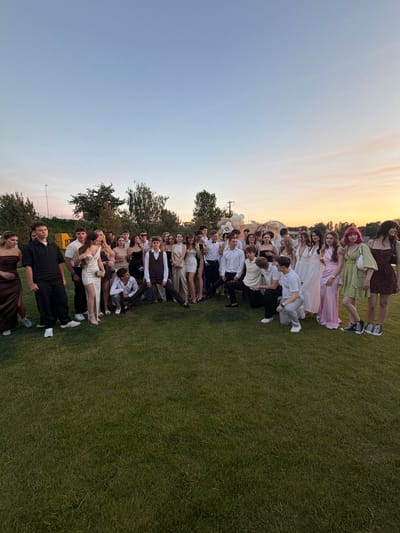 In a grassy outdoor setting, a group of young people are lined up, possibly for a formal event or a photoshoot. The image is taken in Khotianivka, Ukraine. The time of day appears to be late afternoon or early evening, as indicated by the soft, warm light of the setting sun casting a gentle glow across the sky. The weather seems pleasant, with clear skies and ample daylight.

The subjects are dressed in a mix of formal and semi-formal attire. Many are wearing dresses and suits, suggesting a special occasion like a graduation, prom, or wedding. Some individuals have bright pink hair and others are in light-colored dresses, adding pops of color to the scene. One person is holding a bouquet of white balloons. The people are arranged in a staggered line, with some closer to the camera and others further away, creating a sense of depth. Their expressions are varied, with some smiling and others looking more neutral.

The background consists of a vast expanse of green grass, with trees and foliage visible in the distance. The soft sky with wispy clouds adds to the serene atmosphere. A yellow structure, possibly a building or vehicle, is visible behind some of the individuals. The overall impression is one of youthful celebration and camaraderie, captured during a memorable event. There is no visible text in the image.
