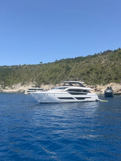 This image captures a scene in Magazia, Greece, featuring a fleet of luxury yachts anchored in the clear blue waters near a lush, green, and rocky coastline. The primary focus is a sleek, white motor yacht with multiple decks and dark-tinted windows, positioned prominently in the foreground. To its left, a second, smaller yacht is visible, appearing mostly submerged except for its upper structure. Further to the right, a dark-colored yacht with a distinctive silhouette rests in the background. The coastline is characterized by steep, verdant slopes dotted with trees and shrubs, with exposed rocky outcrops. The sky above is a vibrant, cloudless blue, indicating a sunny day. The water exhibits gentle ripples, reflecting the bright daylight. There are no people visible in the image.