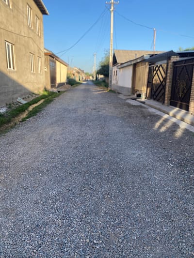 The image depicts a gravel road in a residential area of Боғистон, Tajikistan. The road is lined with houses and fences on both sides. On the left, a two-story building with a stucco facade and multiple windows is visible. A shallow ditch with some grass runs alongside this building. On the right, several single-story houses with corrugated roofs and various types of fences are present, including a dark metal gate with ornate detailing. Utility poles and overhead wires are present, indicating the presence of electricity and communication infrastructure. The sky is clear and blue, suggesting it is daytime, likely morning or late afternoon due to the shadows cast by the buildings. The overall scene conveys a quiet, rural or suburban residential atmosphere. There are no people visible in the image.