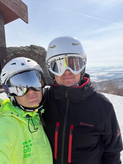 Two individuals, an adult male and an adult female, are depicted outdoors in a snowy, mountainous environment at a ski resort in Zelenaya Polyana, Russia. Both subjects are wearing white ski helmets and mirrored goggles. The female, positioned on the left, wears a neon yellow ski jacket with a partially visible green logo. Her white helmet is branded "ATOMIC." The male, positioned on the right, wears a black ski jacket featuring red zipper accents and a "GLISSADE" logo; his white helmet is branded "WED'ZE." They are in close proximity, facing the camera, likely posing for a selfie. The background displays snow-covered terrain, distant snow-capped hills, exposed rock formations, and a partial view of a dark brown wooden structure on the far left. The sky is clear blue with a visible contrail. The scene indicates a bright, sunny day on a ski slope.