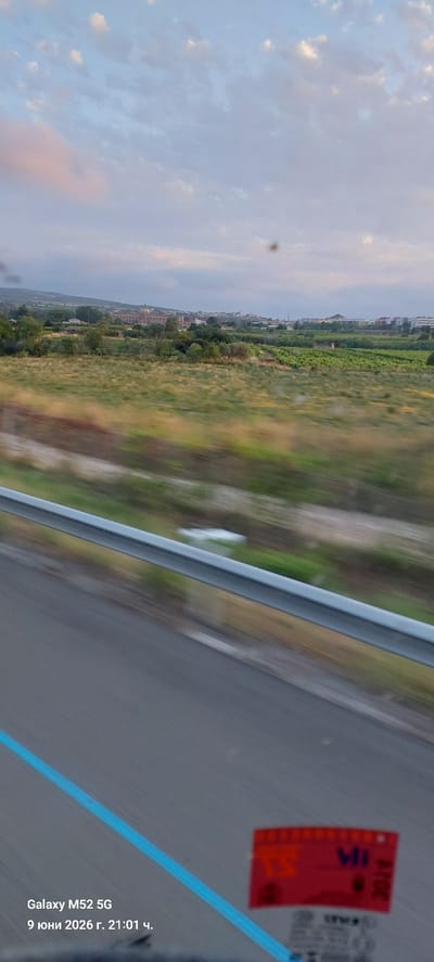 The image depicts a landscape scene viewed from a moving vehicle, likely a train or car, as evidenced by the motion blur. The foreground shows a grey asphalt road with a bright blue line marking the edge, and a silver metal guardrail. A red sticker with white lettering is visible on the right side of the road. In the middle ground, there is a blurred grassy field with some vegetation. The background features a panoramic view of a town or city with buildings and greenery, set against a sky with clouds. The date and time "9 June 2026, 21:01" and "Galaxy M52 5G" are overlaid in the bottom left corner.
The town depicted in the background is Vilafranca del Penedès, Spain. The prominent building visible in the mid-background, to the left of the center, is the Basilica of Santa Maria. The vineyards characteristic of the Penedès wine region are also visible.