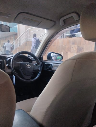 The image captures a view from inside a car, looking out through the front passenger side window and windshield. The car's interior features a beige fabric passenger seat in the foreground, with a dark gray armrest and a partial view of the black dashboard and a steering wheel with a visible Toyota logo. A sun visor is lowered above the windshield, and a beige grab handle is affixed to the ceiling.
Outside the car, two men are visible on concrete stairs with light-colored metal railings, suggesting an urban or semi-urban setting in Sheywe ward, Kenya. The man in the mid-ground, seen from behind, wears a light-colored, possibly plaid shirt and carries a black backpack as he ascends the stairs. Another man, wearing a white t-shirt, is partially visible walking up the stairs behind him. The stairs are built against a wall with a textured, stone-like finish.
The car's side mirror on the right reflects a street scene, showing glimpses of buildings or market stalls with green awnings, indicating a lively area. The natural light suggests it is daytime. No specific weather conditions are discernible, but the overall brightness implies clear skies. The activity depicted is people moving on foot up a set of stairs. No legible text is visible other than the Toyota logo on the steering wheel.