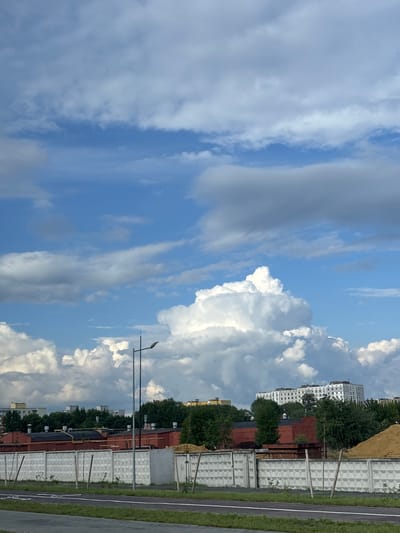 A vertical, eye-level, medium shot captures a cloudy sky over a landscape. The sky is filled with a mix of white, puffy cumulus clouds and wispy cirrus clouds against a bright blue backdrop. In the middle ground, a street light stands tall on the right side of the frame, casting a shadow. Behind it, a long, red brick building with a dark roof is visible, flanked by a line of trees. Further in the background, a multi-story apartment building with numerous windows is seen, also surrounded by trees. The foreground features a paved road and a concrete wall with a repeating diamond pattern, separating it from the scene behind. A pile of sand is visible on the far right side of the image.