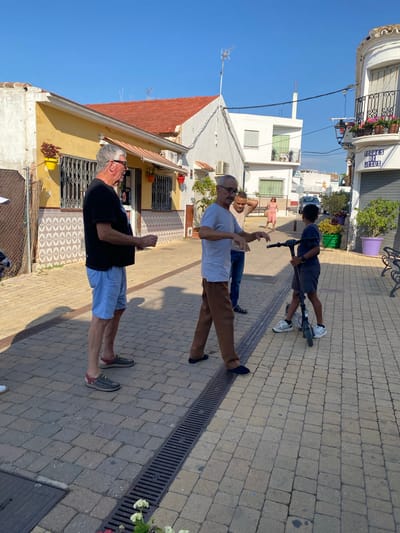The image depicts a street scene in Cancelada, Spain, under a clear blue sky. In the foreground, two adult men and a young boy are interacting. The man on the left, wearing a black t-shirt and light blue shorts, appears to be conversing. To his right, another man in a grey t-shirt and brown pants is gesturing with his hands, seemingly engaged in a discussion with the boy. The boy, dressed in a dark t-shirt and shorts, stands beside a black electric scooter. A building with a yellow facade and red-tiled roof is visible on the left, featuring barred windows and potted plants. Across the street, another building displays a sign that reads "TORRES DE JESUS". The street is paved with cobblestones, and a drainage grate runs along the ground. In the background, additional buildings and vegetation are present, suggesting a residential or commercial area.
