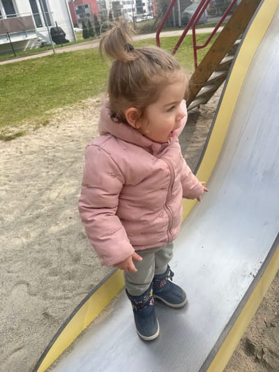 The scene depicts a playground in the city of Senden, Germany, under daylight conditions. A single toddler, estimated to be between 18 months and 3 years old, is positioned on a metal playground slide. The child has light brown hair tied in a high bun, wears small stud earrings, and is dressed in a pink hooded puffer jacket, light grey pants, and dark blue winter boots with colorful motifs. The child is standing upright on the textured surface of the slide, which has yellow side rails, and is looking towards the viewer's left with an open mouth. Their right arm is extended, with the index finger pointing downwards towards the slide's surface. The ground immediately surrounding the slide consists of sandy material. In the background, a grassy area is visible to the left, beyond which modern multi-story residential buildings with balconies are present. To the right of the child, a wooden structure with red metal rails, likely part of the slide's access stairs, is partially visible.