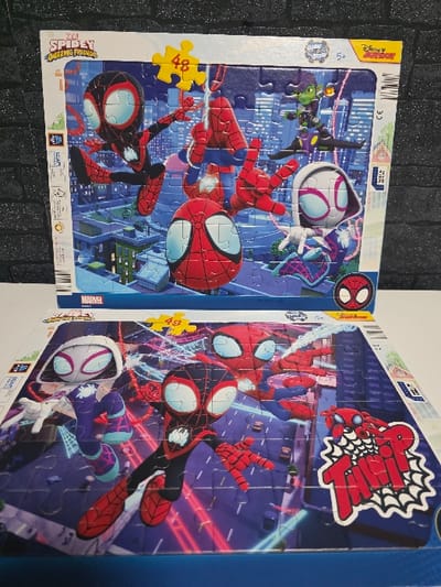 The image displays two partially assembled jigsaw puzzles, stacked one above the other, both featuring characters from "Marvel's Spidey and His Amazing Friends." The items are situated against a dark, textured background resembling a brick wall on the left.

The top puzzle, predominantly visible, depicts four animated characters within a stylized cityscape at dusk. The characters include Spider-Man (classic red and blue suit), Miles Morales (black and red suit), Ghost-Spider (white and purple suit), and Green Goblin (green suit with a purple glider). Spider-Man is positioned centrally, reaching outwards. Miles Morales is to the left, and Ghost-Spider is to the right. Green Goblin is in the upper right, flying on his glider. The cityscape background features various illuminated buildings. A yellow puzzle piece bearing the number "48" is placed in the upper left quadrant. Branding elements visible include "Marvel Spidey and His Amazing Friends," "Glow in the dark," "5+," and "Disney Junior." The puzzle is incomplete, with several individual pieces visible.

The bottom puzzle is mostly assembled and positioned beneath the top one, displaying a different cityscape scene, primarily at night with prominent neon pink and blue light trails. This puzzle features Miles Morales in the foreground, facing right, with Ghost-Spider to his left, and classic Spider-Man to his right. All characters are in dynamic, action-oriented poses. A yellow puzzle piece with the number "48" is in the upper left, and a graphic of a "Thwip!" speech bubble is in the lower right corner. The packaging suggests a 48-piece count for both puzzles.

There are no discernible visual cues or text within the image that link the depicted scenes or the objects themselves to a specific location within the city of Zweibrücken, Germany. The backgrounds are generic, animated cityscapes.