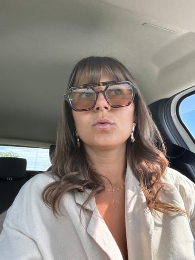 A woman with brown hair and bangs is wearing tortoiseshell-framed glasses and a light-colored linen blouse. She is wearing gold earrings and a delicate pearl necklace. She is sitting in a car, as evidenced by the headrest, window, seatbelt, and interior car panels. The location cannot be identified as Saint-Cyprien, France, based on the image content alone.