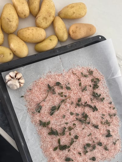A top-down photographic view captures a food preparation scene. The upper portion of the frame features approximately fifteen to twenty unpeeled, whole, light-yellow potatoes scattered on a light-colored countertop. A black baking sheet, lined with white parchment paper, occupies the lower section of the image. On the parchment paper, a substantial quantity of coarse, light-pink granular salt, identified as Himalayan pink salt, forms a mound, primarily towards the right. Numerous small, green herb sprigs and individual leaves, resembling oregano or thyme, are distributed across the salt. To the left of the salt, on the parchment paper, rests a whole head of garlic, slightly broken to reveal its cloves. The lighting is bright and uniform. The scene is located in the city of Ariel, Palestinian Territories.