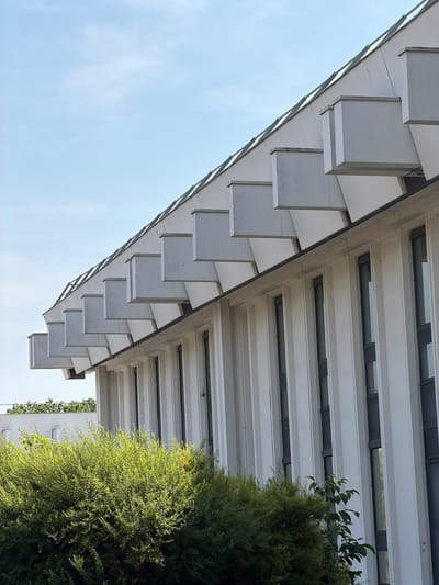 The image shows a section of a modern white building with a distinctive roofline featuring repeating geometric protrusions. The building has a facade with vertical white panels and dark-framed windows arranged in a repeating pattern. In the foreground, a dense, green bush is visible. The sky above is a clear blue with a few faint clouds. In the background, another white building can be seen, partially obscured by trees. The architectural style suggests it could be a public building, possibly an institutional or administrative one. The location is identified as Rungis, France, a commune known for its wholesale market and a location with a mix of commercial and residential architecture. The image captures a bright, sunny day, likely outdoors. There are no people or actions depicted in the scene.