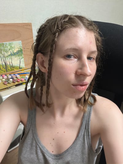 A young Caucasian woman with light skin and light brown hair styled in multiple braids is pictured in a selfie. She is wearing a grey tank top. Her expression is neutral, and she is looking towards the camera. In the background, an easel holds a painting and art supplies, including small pots of paint and brushes. The setting appears to be an indoor space, possibly an art studio or a room within a residence. The image does not contain any information that would allow for the determination of its location in Dongducheon-si, South Korea.