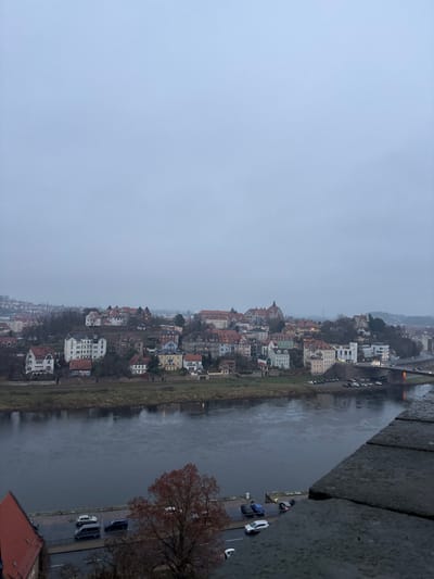 The image presents a panoramic view of the town of Meissen, Germany, situated along a river, under an overcast sky. The viewpoint is from an elevated position, likely a bridge or a high structure, looking down and across the river.
A significant portion of the image, particularly the upper left, is filled with a uniform, light grey sky, indicating dull or overcast weather conditions. A wide river flows through the scene, from left to right, reflecting the muted light.
Along the far bank of the river, the town's buildings are densely packed, extending along the waterway and upwards onto a gentle slope. The architecture is distinctly European, with numerous multi-story buildings displaying a variety of colors and prominent pitched roofs, many of which are reddish-brown. On the closer bank, a road or embankment runs parallel to the river, with several cars parked along its side. A large, bare tree with reddish-brown leaves stands out in the lower right foreground. In the upper right corner, part of a bridge or elevated road structure crosses the river, with faint vehicle lights visible.
The immediate foreground on the right includes a rugged stone wall or parapet, suggesting the viewing platform, and partially visible below it, a red-tiled roof. The scene depicts a tranquil, everyday view of a town. The bare trees and grey sky suggest a cooler season, possibly late autumn or winter. There are no specific human activities or individuals prominently featured, and no visible text is present.