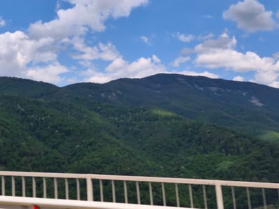 The image depicts a scenic view of a mountainous landscape, likely taken from a bridge or elevated vantage point. The foreground features a white metal railing, suggesting the photographer is on a structure crossing a valley or overpass. Behind the railing, rolling green hills covered in dense forest dominate the scene. The trees appear lush and vibrant, indicating a time of year with active vegetation growth, possibly late spring or summer.
In the background, a larger, darker mountain range rises, its slopes also covered in trees but with some lighter, rocky patches visible on its upper reaches. The sky above is a bright blue, dotted with fluffy white cumulus clouds. Sunlight filters through the clouds, creating areas of bright illumination and shadow on the mountainside, adding depth and texture to the landscape. One of the cropped images shows a tiny speck in the sky that could be an airplane or bird.
The overall impression is one of natural beauty and tranquility. There is no visible text in the image, and no people or specific objects are identifiable besides the railing and the natural elements of the landscape. The time of day appears to be daytime, with the sun casting distinct shadows, and the weather seems pleasant and clear, with scattered clouds. The location context provided places the scene in Vrachesh, Bulgaria, a region known for its mountainous terrain and natural beauty.