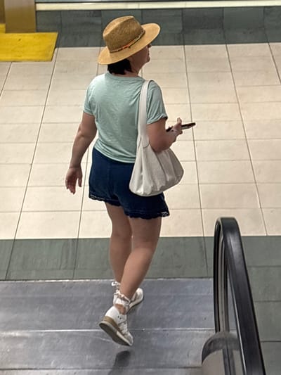 A person wearing a straw hat, a light blue t-shirt, and dark blue shorts is standing on an escalator. They are holding a smartphone in their right hand and a white purse is slung over their left shoulder. The person is wearing white sneakers with brown accents. The background appears to be a tiled wall and the metal railing of the escalator. The lighting suggests it could be daytime, and the setting seems to be an indoor public space like a shopping mall or train station. There are no visible emotions or text in the image.
