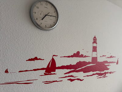 A circular clock with a silver frame hangs on a textured white wall. The clock face is white with black numerals and hands. A smaller dial for a hygrometer and a thermometer is located at the top of the clock face. Below the clock, a red sticker mural depicts a nautical scene. The mural includes a lighthouse on a rocky outcrop, two sailboats on the water, and several stylized clouds. A small bird is also depicted in the mural. The image was taken in the city of Fehmarn, Germany.