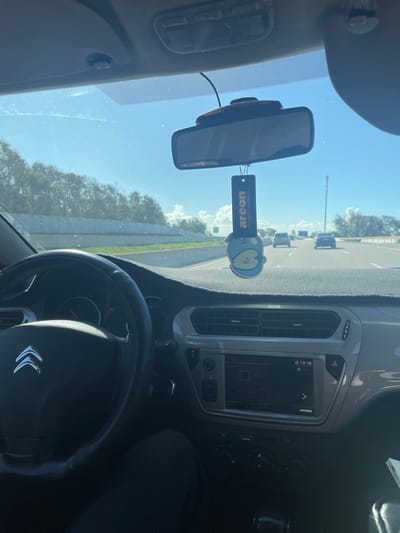 The image depicts an interior view from a Citroën vehicle, positioned as if from the driver's seat, looking forward onto a multi-lane highway near Skhirat, Morocco, during daylight hours. A driver's left arm and leg, clad in dark clothing, are partially visible. The black steering wheel, featuring a silver Citroën emblem, is prominent on the left. The grey dashboard extends across the foreground, partially covered by a dark, textured mat. A central infotainment system displays the time as "13:18" and "Radio" mode, flanked by air vents and climate control knobs. A rearview mirror is mounted centrally above the dashboard, with a small electronic device attached above it. Suspended from the rearview mirror is a blue, cartoon-character shaped air freshener bearing the "Areon" brand.
Through the windshield, the highway, marked by clear asphalt, stretches into the distance, with three other vehicles visible ahead, traveling in the same direction. To the left of the highway, a concrete barrier separates the road from a dense line of green trees. To the right, a wide shoulder or median leads to additional vegetation and a tall, slender pole. The sky above is clear blue with scattered white cumulus clouds, indicating bright, sunny weather.
