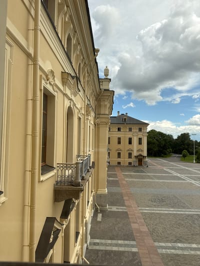 The image shows a side view of a pale yellow building with ornate architectural details, including decorative window surrounds and a balcony with an iron railing. The building features tall, arched windows and is partially obscured by a vertical drainpipe. In the background, across a paved courtyard with a reddish-brown pathway, stands another pale yellow building with a dark roof and multiple windows. Beyond this second building, a line of green trees forms a backdrop, and a cloudy sky with patches of blue is visible above. The overall scene suggests an exterior view of a historical or institutional building, likely within a public square or park in Strelna, Russia. No people are visible in the image.