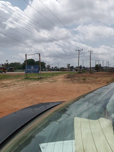 The image depicts an outdoor scene under a cloudy sky, likely during the day. In the foreground, the back of a car is visible, with its rear windshield reflecting the surrounding environment. The scene shows a dirt road leading to a paved road, with several electrical poles and power lines crisscrossing the sky.
Further in the background, there is a blue sign with white lettering that reads "FEDERAL ROAD SAFETY CORPS PLATEAU STATE SECTOR COMMAND RS4.1 KM ZARIA ROAD, JOS PLATEAU STATE." This indicates the location is within Plateau State, Nigeria, specifically on Zaria Road in Jos.
To the left of the sign, a green sign is partially visible, which appears to say "FEDERAL GOVERNMENT COLLEGE". There are also a few vehicles on the paved road, including what looks like a tricycle, a common mode of transport in Nigeria. People are seen walking and standing near the road.
The overall setting suggests a roadside view in a semi-urban or rural area of Nigeria. The weather appears overcast, with significant cloud cover. The presence of the Federal Road Safety Corps sign points to an official government presence and a focus on road safety. The image captures a slice of everyday life on a Nigerian road.
