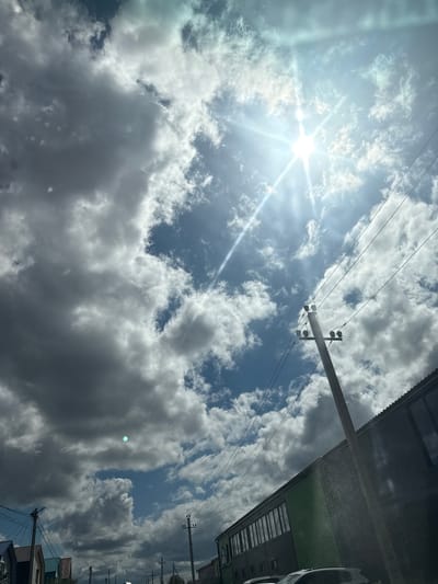 This is a photograph taken from a low-angle perspective, looking upwards at a dynamic sky filled with cumulus clouds. The sun is bright and appears to be high in the sky, with visible lens flares and rays of light streaming through gaps in the clouds. The sun's intensity suggests it is midday. The clouds range from bright white, where they are catching the sun's direct light, to dark gray, indicating depth and thickness.

In the foreground and mid-ground, several elements suggest an urban or semi-urban environment. A utility pole with insulators and wires is prominent on the right side of the image, stretching diagonally upwards. More power lines crisscross the sky. Partially visible on the right edge is the roofline of a building, appearing to have a corrugated metal surface and a green section. Further down the building, architectural details like windows and a roof are visible, with a hint of a car parked nearby. The overall scene is one of a bright, possibly warm day with a mix of sunshine and cloud cover. There is no visible text in the image. The angle and composition give a sense of looking up from street level, possibly from a car or a sidewalk, towards the sky and the overhead infrastructure.