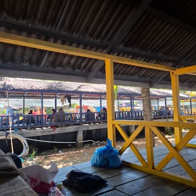 The image depicts an outdoor covered seating area, likely a restaurant or rest stop, situated along a waterfront in Benteng, Indonesia. The structure features a corrugated metal roof supported by dark beams, with a yellow painted wooden railing in the foreground. Beneath the roof, a blue painted railing separates the seating area from a walkway or pier. Several individuals are seated along the blue railing, some facing away from the camera. Clothing items, including what appears to be a red jacket and a dark t-shirt, are visible on the walkway. A large blue plastic bag, presumably containing trash, sits on the wooden floorboards in the foreground, next to a pile of folded dark clothing and a partially visible box of tissues. The ground beneath the structure is sandy and littered with debris, including plastic bottles and scattered trash. The water is not clearly visible, but the context suggests a canal or riverbank setting. The overall impression is one of a functional, somewhat weathered, public space.