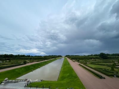 The image shows a view of the Peterhof Palace complex in Strelna, Russia, on a cloudy day. In the foreground, there are stone stairs leading down to a canal or water channel. The canal is long and rectangular, with grass growing on its banks. On either side of the canal are meticulously maintained gardens with manicured hedges and pathways. In the distance, the canal leads to a building that appears to be part of the palace. The sky is filled with dramatic, dark clouds, suggesting an approaching storm. There are no visible people in the image, only the architectural and landscape elements of the palace grounds.
