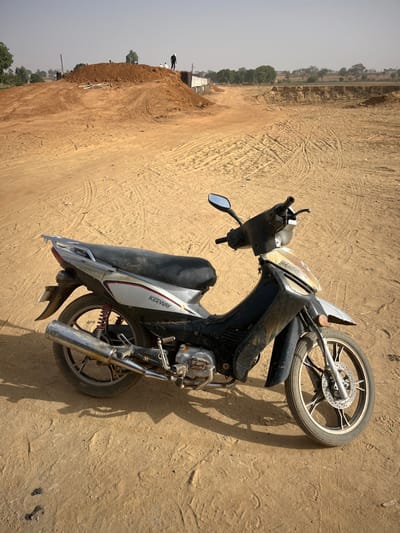 A Keeway motorcycle lies sideways on a dusty, dirt-covered ground. The vehicle is primarily black with silver accents. Its front wheel is visible, casting a shadow. In the background, a beige landscape stretches with a small green area on the horizon, possible trees and some buildings under construction. The sky is slightly hazy.