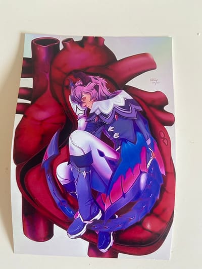 The image shows a printed illustration of a stylized anime character with pink hair, a purple and blue outfit, and white leggings, curled up inside a large, anatomically detailed red heart. The heart appears to be depicted as a visceral organ with its chambers and major blood vessels. The character is resting their head on their arm, with eyes closed. In the upper right corner of the illustration, there is a small signature that reads "Nelly 20/23". The print is placed on a white surface, possibly a table or wall, with some slight shadows and variations in lighting. There is no visible context or connection to the city of Kassel, Germany.