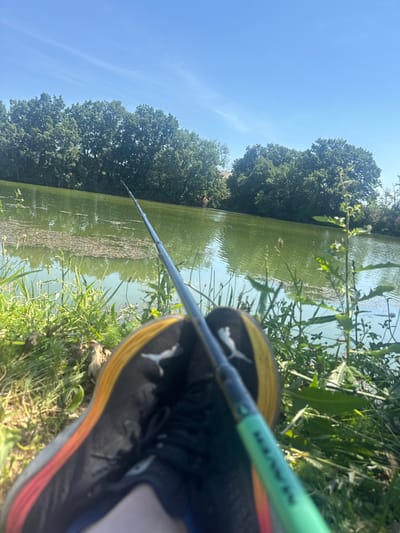 The image depicts a first-person perspective of someone fishing by a body of water on a sunny day. The foreground shows a black athletic shoe with orange and yellow accents and a white logo, placed on grassy ground. A fishing rod, predominantly green with some black sections, extends into the frame from the shoe and over the water. The water, appearing murky green, is partially covered with debris and surrounded by lush green vegetation and trees along the bank. The sky is clear blue. The scene appears to be outdoors in a natural setting, likely a pond or small lake. No specific location in Smilets, Bulgaria can be identified from the visual information alone.