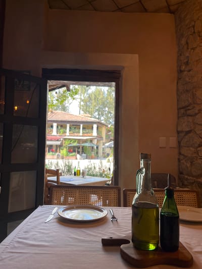 The image captures a dining scene in what appears to be a restaurant or similar establishment, situated in the city of Domingo Arenas, Mexico. The view through the window reveals a multi-story building with architectural features typical of Mexican design, complete with balconies, terracotta roofs, and visible greenery. Inside, the focus is on a table setting adorned with a light-colored tablecloth. Place settings include plates and cutlery. Prominently featured are two glass bottles containing liquids, placed on a wooden serving board. The lighting is soft and diffused. The overall ambiance suggests a quiet, possibly daytime, setting. The physical environment includes stone walls and furniture with woven elements. No people are visible in the shot.